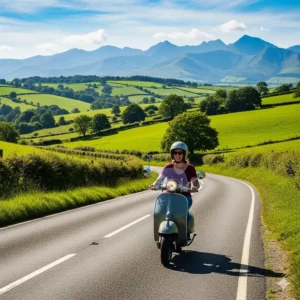 A woman drive scooter on a winding road through the countryside, with green fields and distant mountains visible under a clear blue sky.